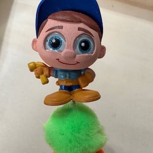 May include: A blue and brown figurine of a construction worker wearing a blue baseball cap and holding a yellow hammer. The figurine is standing on top of a stack of three pom-poms, one green, one orange, and one yellow.