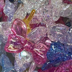 May include: A close-up shot of a pile of iridescent, clear, and colorful glass bow-shaped beads. The beads are in shades of pink, purple, blue, and yellow. The light reflects off the surfaces, creating a sparkling effect. These beads are suitable for jewelry making and crafts.