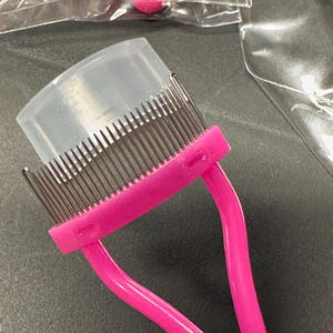 May include: A pink eyelash comb with a clear plastic cover. The comb has thin metal teeth and a curved handle. The tool is designed for separating and grooming eyelashes. The background is a dark gray surface.