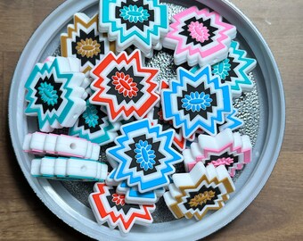 May include: A collection of colorful, geometric patterned silicone charms in a variety of colors, including blue, red, pink, gold, and white. The charms are shaped like stars and have a raised design.
