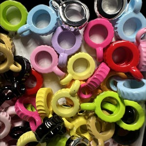 May include: A collection of colorful, ring-shaped objects with a small handle. The rings come in various colors, including pink, blue, green, yellow, black, and silver. The objects appear to be made of plastic or a similar material.