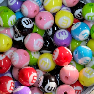 May include: A collection of colorful plastic beads with the letter "m" printed on them. The beads are in various colors, including pink, yellow, green, blue, red, purple, and black.
