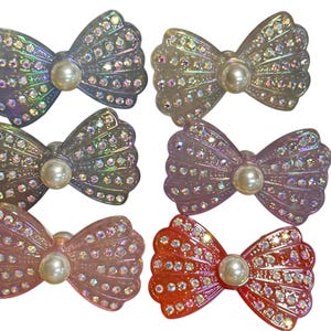May include: Six decorative bow-shaped hair clips in various colors, including iridescent, pink, and orange. Each clip features a pearl center and is embellished with small, sparkling rhinestones. These accessories are ideal for adding a touch of glamour to any hairstyle.