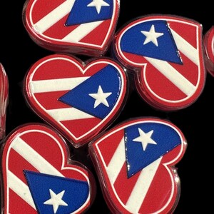 May include: A collection of heart-shaped charms featuring the Puerto Rican flag design. Each charm displays a red and white striped pattern with a blue triangle on the left side containing a single white star, all finished with a glossy surface.
