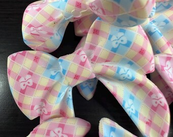 May include: A collection of pastel-colored fabric bows. Each bow features a checkered pattern in pink, yellow, and blue, with small floral accents. The bows are arranged in a close-up shot, showcasing their delicate design and texture.