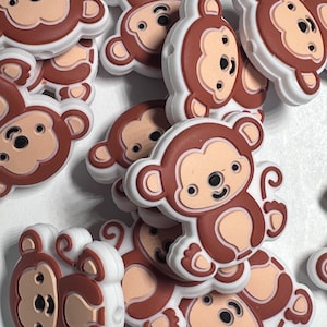 May include: A collection of brown, tan, and white monkey-shaped charms. Each charm features a smiling monkey face with black eyes and a tan face, surrounded by a white border. The charms are scattered across a white surface.