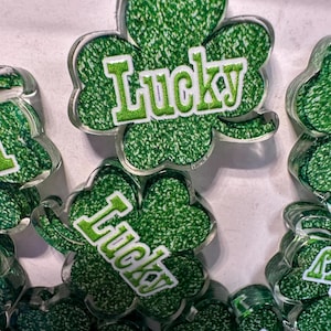 May include: Clear acrylic shamrock-shaped charms with a green glitter fill and the word "Lucky" in white and green. These charms are ideal for St. Patrick's Day crafts and decorations.