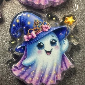 May include: A clear acrylic charm featuring a cartoon ghost wearing a blue witch hat with a gold crown and pink bows. The ghost has a smiling face, rosy cheeks, and holds a yellow star-tipped wand. The design includes purple and blue gradients.