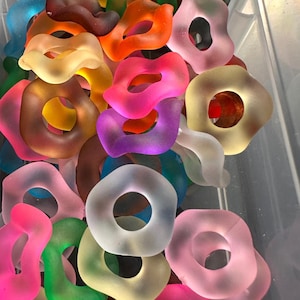 May include: A collection of colorful, translucent, ring-shaped glass beads in various shades including pink, orange, yellow, green, and purple. The beads have a smooth, frosted appearance and are arranged closely together.