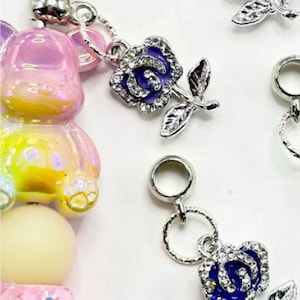 May include: Close-up of jewelry charms featuring a blue rose design with silver-colored leaves and a pink rose design. The charms are attached to a beaded accessory with pink, yellow, and iridescent beads.