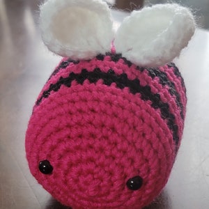 May include: A pink and black crocheted bee with white crocheted wings. The bee has black eyes and a striped body.