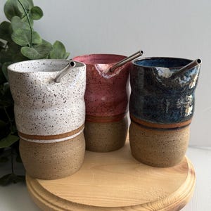 Puede incluir: Tres tazas de cer&aacute;mica con esmalte marr&oacute;n moteado y una banda marr&oacute;n alrededor de la parte superior. Cada taza tiene una pajita de acero inoxidable y es de un color diferente: blanco, rosa y azul.