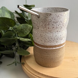 Puede incluir: Una taza de cer&aacute;mica blanca con motas marrones y una banda marr&oacute;n alrededor de la parte superior. La taza est&aacute; sentada sobre una posavasos de madera y tiene una pajita de acero inoxidable que sobresale.
