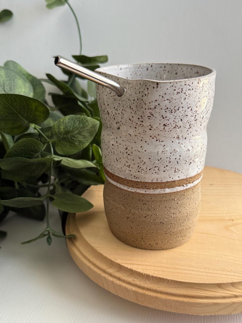 Puede incluir: Una taza de cer&aacute;mica blanca con motas marrones y una banda marr&oacute;n alrededor de la parte superior. La taza est&aacute; sentada sobre una posavasos de madera y tiene una pajita de acero inoxidable que sobresale.