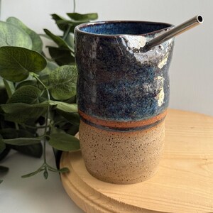 Puede incluir: Una taza de cer&aacute;mica azul y marr&oacute;n con una pajita de metal. La taza tiene una superficie texturizada y una banda marr&oacute;n alrededor del medio. La taza est&aacute; sentada sobre una superficie de madera.