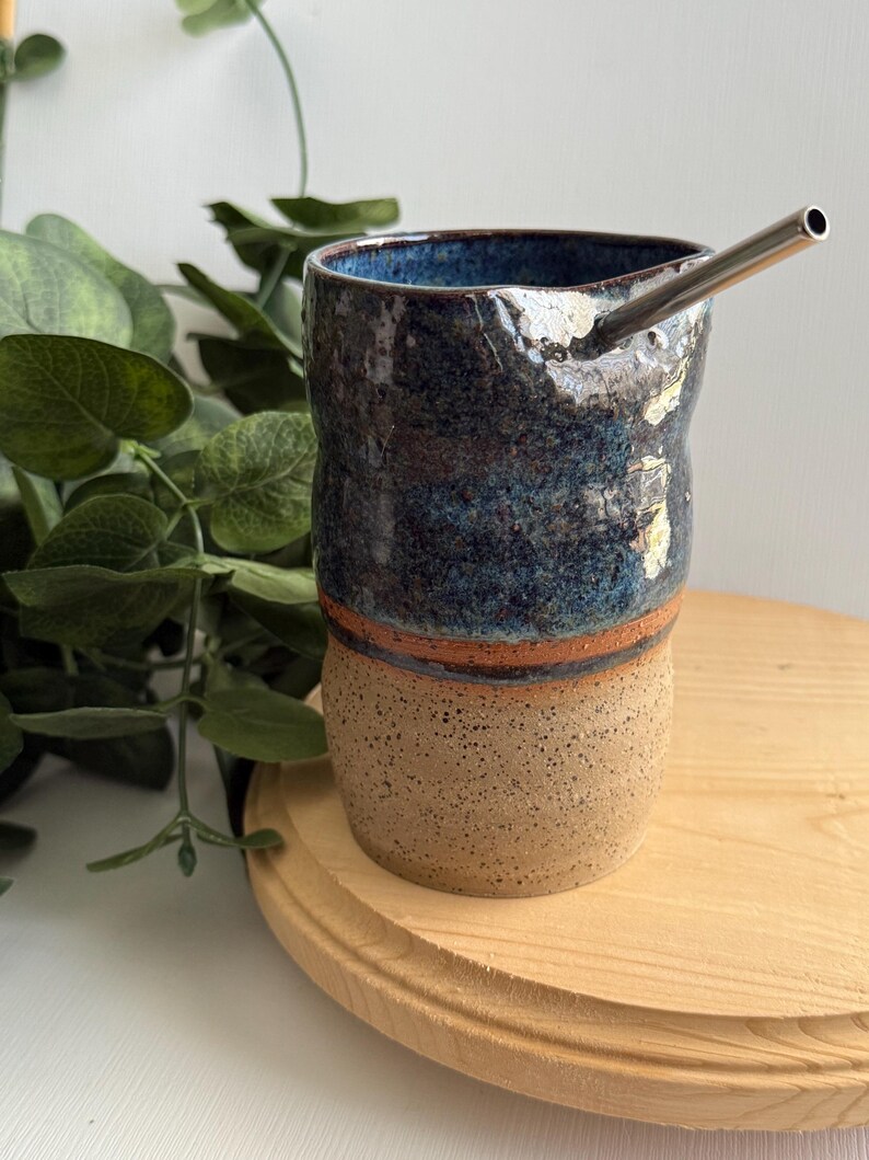 Puede incluir: Una taza de cer&aacute;mica azul y marr&oacute;n con una pajita de metal. La taza tiene una superficie texturizada y una banda marr&oacute;n alrededor del medio. La taza est&aacute; sentada sobre una superficie de madera.