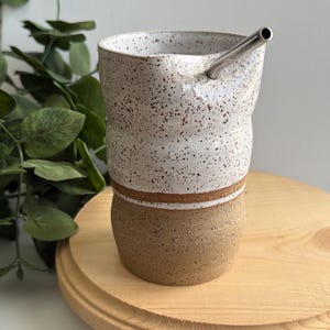 Puede incluir: Una taza de cer&aacute;mica blanca con motas marrones y una franja marr&oacute;n alrededor del medio. La taza est&aacute; sobre una superficie de madera y tiene una pajita de acero inoxidable que sobresale de la parte superior.