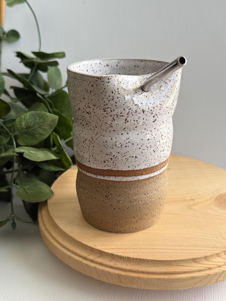 Puede incluir: Una taza de cer&aacute;mica blanca con motas marrones y una franja marr&oacute;n alrededor del medio. La taza est&aacute; sobre una superficie de madera y tiene una pajita de acero inoxidable que sobresale de la parte superior.