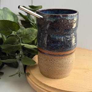 Puede incluir: Una taza de cer&aacute;mica hecha a mano con un esmalte azul y marr&oacute;n. La taza tiene una forma ancha y redondeada y una superficie texturizada. Una pajita de acero inoxidable sobresale de la taza.