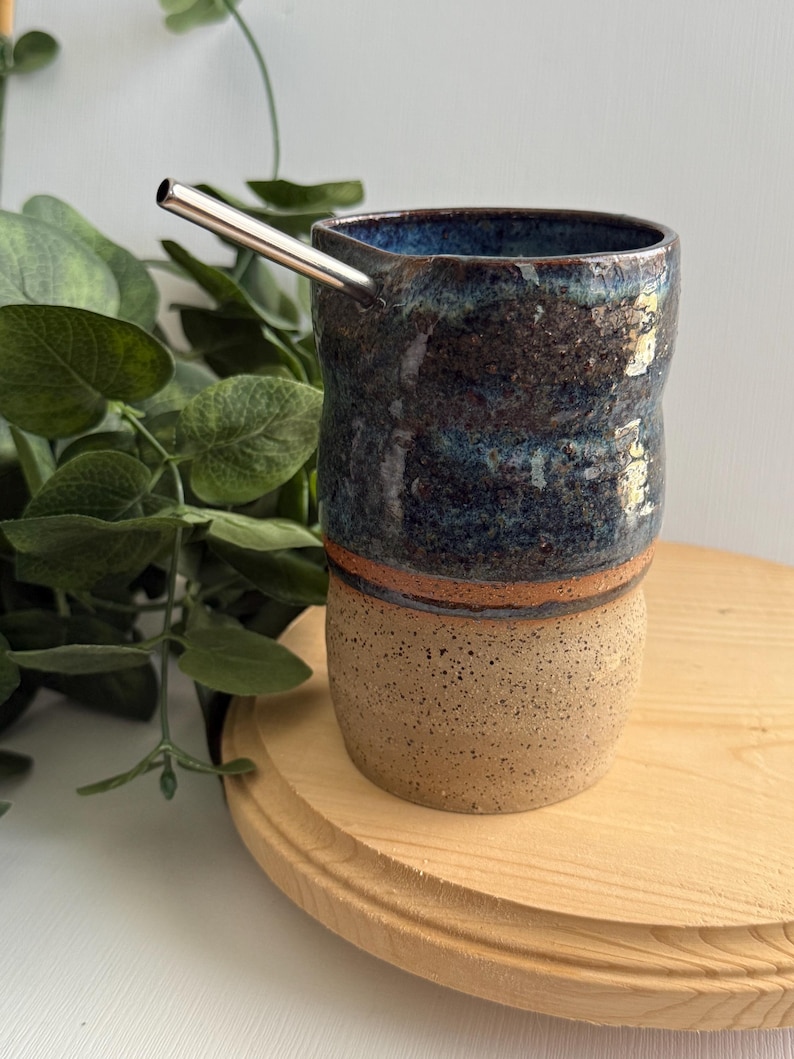 Puede incluir: Una taza de cer&aacute;mica hecha a mano con un esmalte azul y marr&oacute;n. La taza tiene una forma ancha y redondeada y una superficie texturizada. Una pajita de acero inoxidable sobresale de la taza.