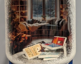 May include: A white glass ornament with a snowy scene inside. The scene shows a cozy living room with a window, a couch, and a bookshelf. There are several books on the floor, including one with the title "The Pact" by Jodi Picoult. The ornament has a blue glitter base.