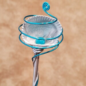 May include: A teal blue wire sculpture of a flower with a white shell-like center and a teal blue bead in the center.