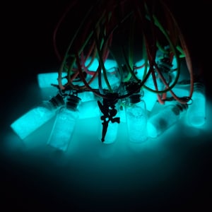 May include: A group of small glass bottles filled with glowing blue glitter, hanging from colorful cords. The bottles are arranged in a cluster, with some overlapping. The glitter inside the bottles is illuminated, creating a bright blue glow.