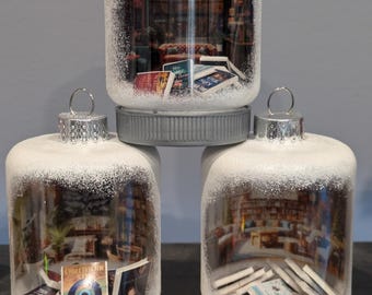May include: Three white glass ornaments with a snowy finish. Each ornament contains a miniature scene of a bookstore interior with books and a small potted plant. The top ornament has a silver lid and the bottom two have silver tops.