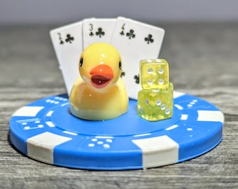 May include: A yellow rubber duck with an orange beak sits on a blue and white poker chip. Two clear dice and three playing cards are also present. The cards show the 2, 3, and 4 of clubs.