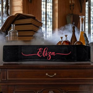 May include: A black rectangular box with the name "Eliza" in pink script. The box sits on a wooden dresser with a drawer. Several glass bottles and stacks of books are in the background, with a smoky effect.
