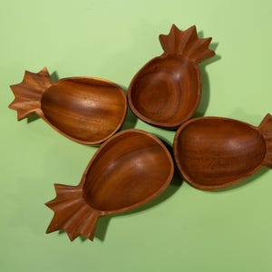 May include: Four wooden pineapple-shaped bowls arranged on a green surface. The bowls are made of a dark brown wood and have a carved, textured design. The bowls are empty and ready to be filled with snacks or other items.