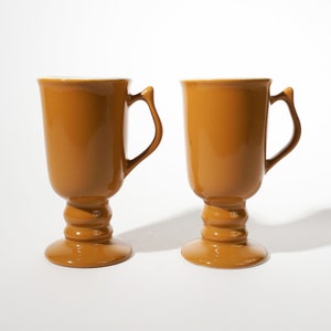 May include: Two brown ceramic mugs with white interiors and a footed base. The mugs have a rounded shape and a handle on the side.