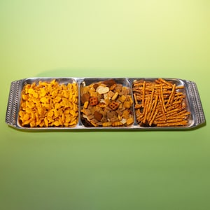 May include: A silver serving tray with three compartments filled with snacks. The left compartment has goldfish crackers, the center has a mix of nuts and crackers, and the right has pretzel sticks. The tray is on a green background.