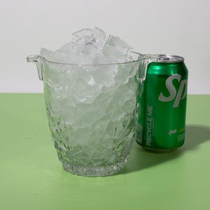 May include: A clear glass ice bucket filled with ice cubes. A green can of soda with the word 