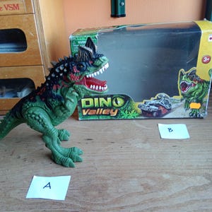 Op de afbeelding: Een groen en zwart dinosaurus speelgoed met rode accenten, open mond en scherpe tanden. Het speelgoed staat naast een doos met de woorden "Dino Valley" en een dinosaurus illustratie. Het speelgoed is ongeveer 25 cm lang.