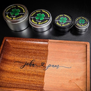 May include: A wooden tray with a dark brown finish on one side and a lighter brown finish on the other side. The tray is engraved with the names "John & Pam". There are four small round containers with labels that say "Lucky One Wood Butter".