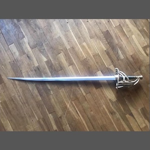 Peut inclure: Une épée argentée avec une garde décorative et une longue lame droite. L'épée est posée sur une surface en bois.