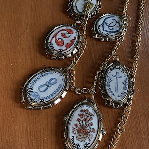 Puede incluir: Un collar vintage dorado con múltiples colgantes ovalados con diseños intrincados. Cada colgante presenta ilustraciones pintadas a mano, incluyendo arreglos florales, cruces y patrones abstractos. El collar tiene una cadena delicada.