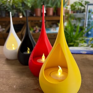May include: Four teardrop-shaped candle holders in white, black, red, and yellow. Each holder features a hollow centre containing a small, battery-operated LED tea light with a warm, glowing flame. The smooth, matte-finished structures taper to a point.