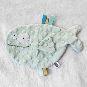 Peut inclure: Un jouet en peluche en forme de poisson bleu clair avec un visage souriant, de grands yeux et une surface texturée. Le poisson a une petite nageoire et une queue. Le jouet est doté de rubans colorés.