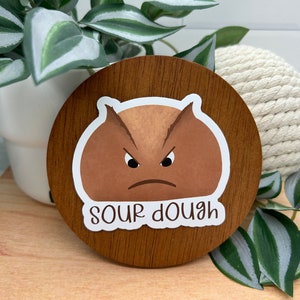 May include: A round wooden coaster with a brown and white sticker of a cartoon sourdough bread with a frowning face and the text "sour dough".