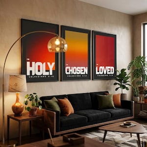 Puede incluir: Una escena de salón con tres impresiones artísticas enmarcadas con las palabras "HOLY", "CHOSEN" y "LOVED". Un sofá negro con cojines coloridos se encuentra debajo de las obras de arte. Una lámpara de arco dorada y una mesa auxiliar de madera con una lámpara completan la decoración.