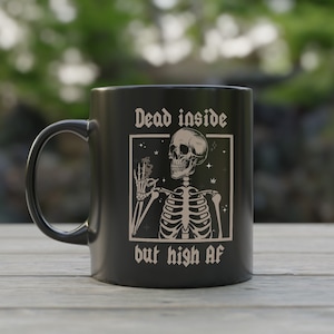 May include: Black ceramic coffee mug with a white graphic of a skeleton smoking a joint. The text on the mug reads "Dead inside but high af."