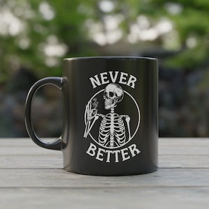 May include: Black ceramic mug with a white graphic of a skeleton inside a circle, with the words "NEVER BETTER" in white. The mug has a curved handle and is set on a wooden surface against a blurred green background.