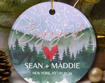 May include: A round, white, ceramic ornament with a watercolor design of a forest and a starry sky. The text "Engaged" is written in cursive in red. Below the text is a red heart and the names "Sean + Maddie" in black. The text "New York, NY | 02.27.24" is written in black below the names.