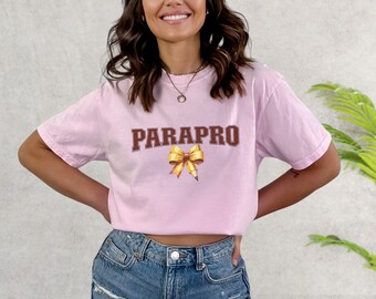 May include: A pink t-shirt with the text "PARAPRO" in brown letters and a yellow bow. The shirt is worn by a person with long brown hair.