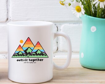 May include: White ceramic mug with a colorful mountain design and the text "outside together" with the tagline "fresh air - quality time". The mug is sitting on a wooden surface next to a light blue pitcher with white polka dots filled with a bouquet of blue and white flowers.