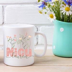May include: White ceramic mug with a pink floral design and the text "NICU Lauren". The mug is sitting on a wooden surface next to a light blue pitcher with white polka dots filled with a bouquet of blue and white flowers.