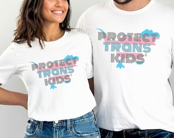 May include: Two white t-shirts with the text "PROTECT TRANS KIDS" in black and blue lettering with a pink and blue design. The shirts are being modeled by two people.
