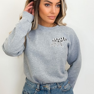 May include: A gray sweatshirt with a black bear silhouette on the front. The bear is made up of the word "MAMA" and other words. The model is wearing the sweatshirt with denim shorts.